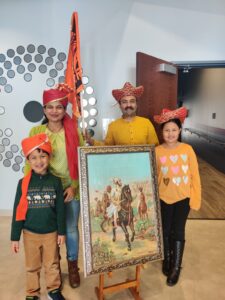 A mother, father, daughter and son dressed in traditional cultural headwear proudly display a flag and painted historical photo in the theater lobby.