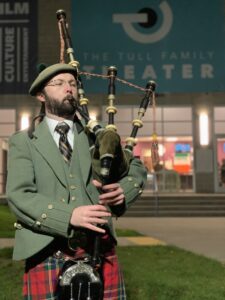 A bearded man wearing traditional Scottish garb holds his lips to the bagpipes he's holding in front of the theater
