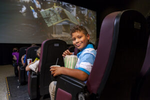 A young boy sitting in a theater chair holding a popcorn and soda gives the camera a smirk as a movie plays on the screen in front of him.